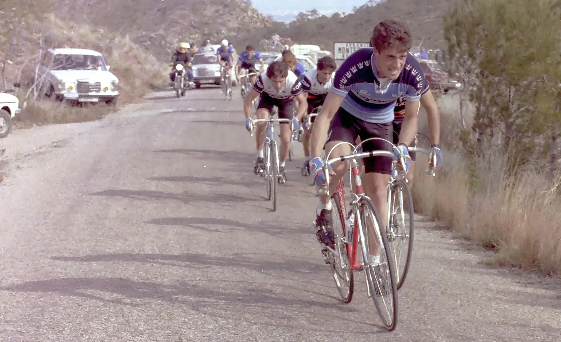Un grupo de corredores sube una carretera de montaña en 1982, con coches de equipo detrás
