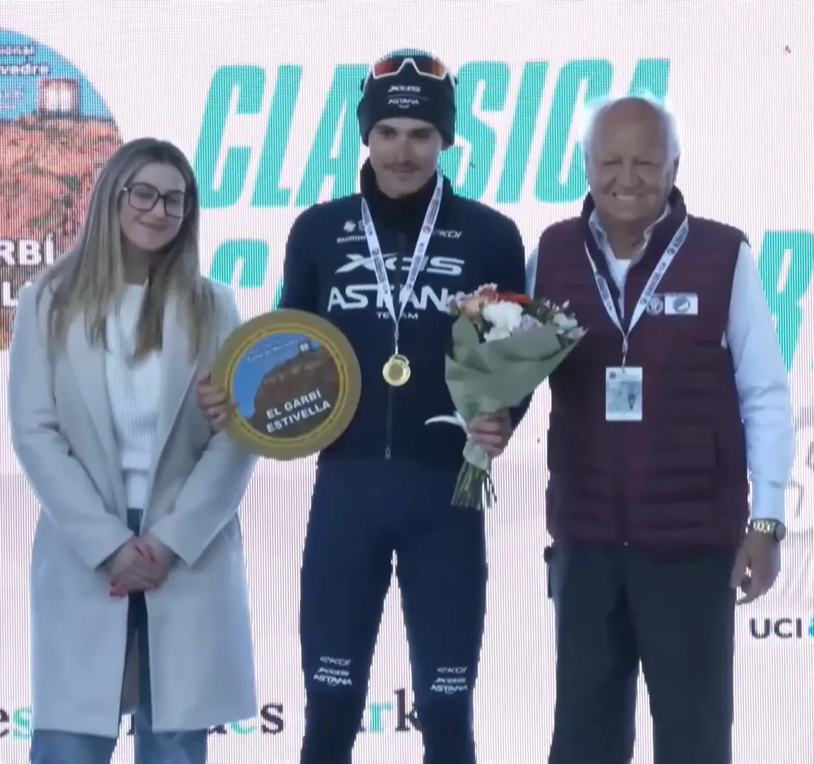 Christian Scaroni, ganador de 2026, sonríe en el podio con el trofeo del Garbí y un ramo de flores, junto a José Ramón Mateu y una persona de la organización